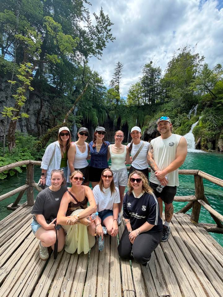 Travellers posing on a wooden bridge with emerald pools, lush forest and cascading waterfalls behind them.