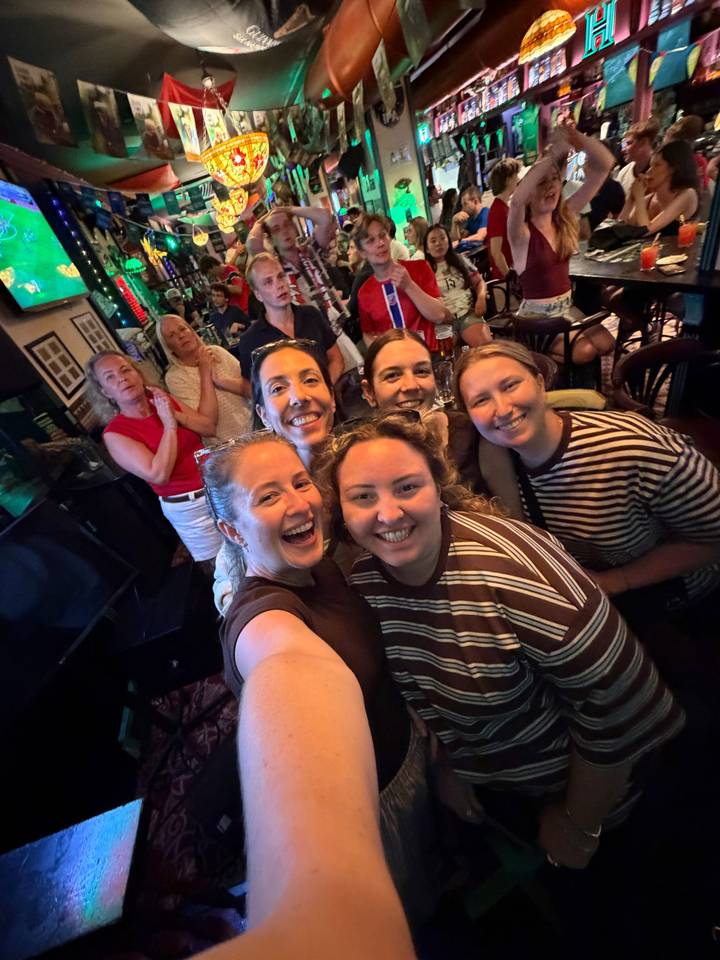 Indoor selfie of friends laughing together in a busy bar with bright screens.