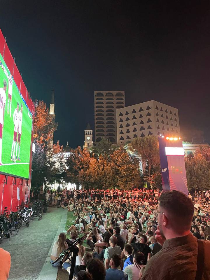 Nighttime public viewing area with a giant outdoor screen showing football in a city square.