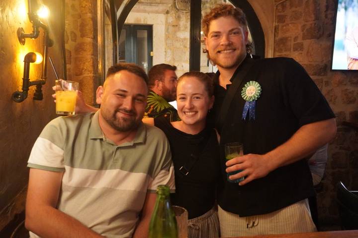 Three friends enjoy drinks and smiles inside a dimly lit stone-walled bar.