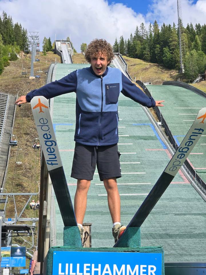 Young man balances with arms outstretched between the uprights of a ski jump ramp overlooking a green landing hill.