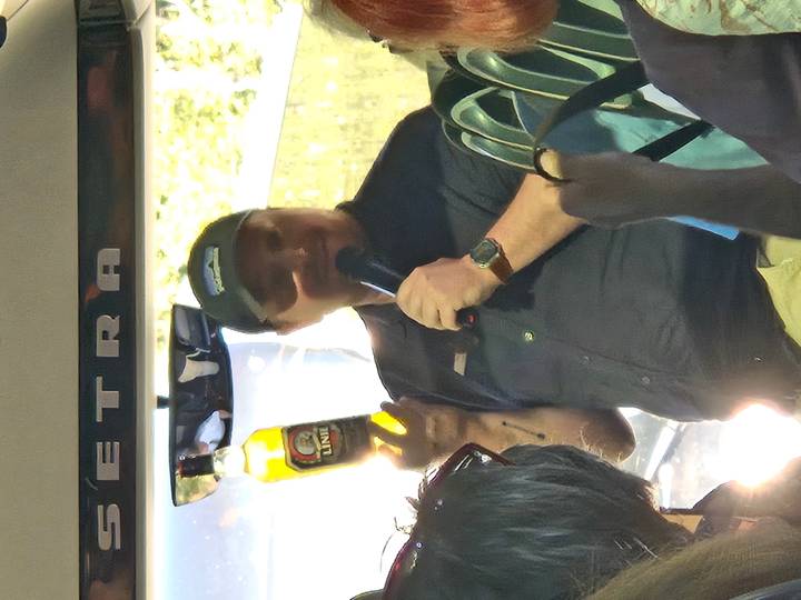 Tour guide on a coach enthusiastically holds up a bottle and speaks into a microphone to passengers.