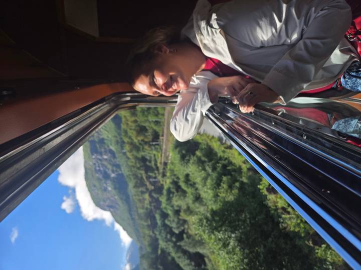 Woman leans out of an open train window, smiling at verdant mountains and valley scenery beneath a blue sky with clouds.