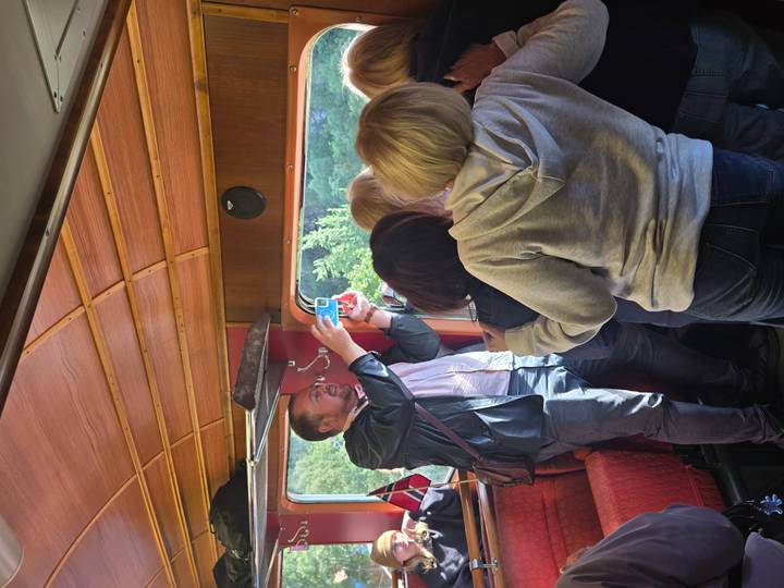 Passengers crowd inside a vintage wooden-ceiling train carriage while a man takes photos toward the window.
