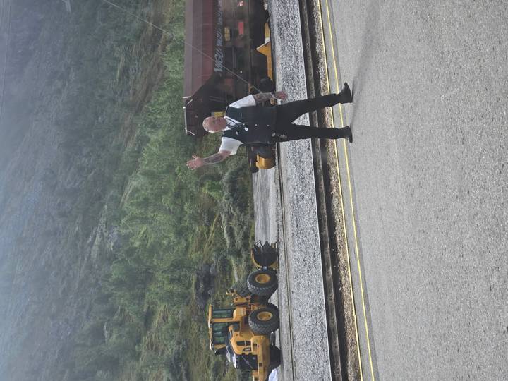Railway staff member in uniform waves from beside the tracks with heavy machinery in the background.