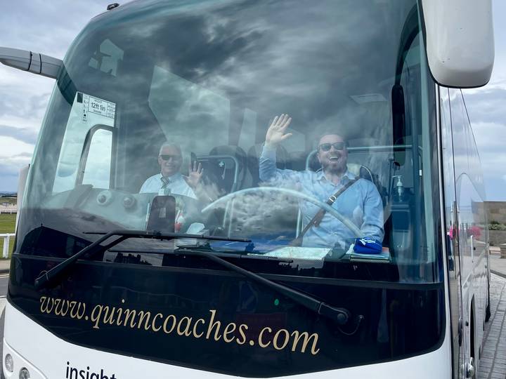 Driver and guide wave cheerfully from the front seats through the large windshield of a tour coach.