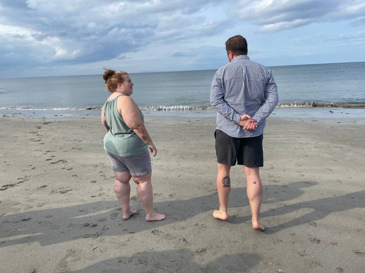 Two people stand barefoot on a quiet grey sand beach looking out over calm sea and sky.