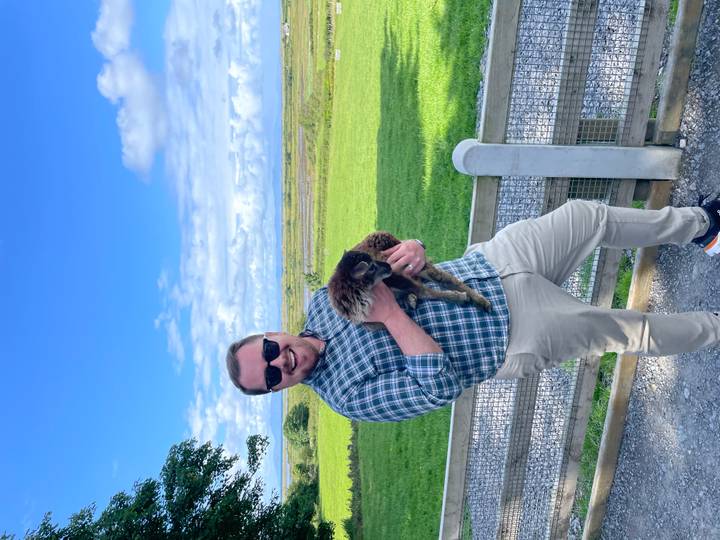 Man smiles while holding a small lamb on a rural farm with green pastures behind.