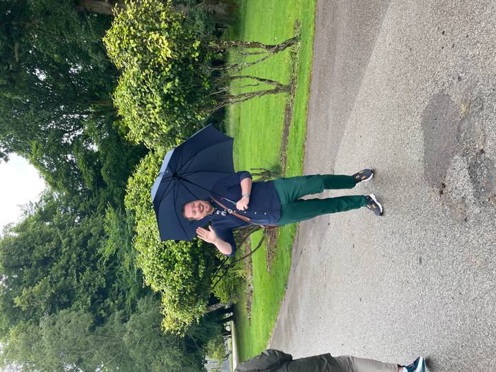 Man waves happily while holding a large umbrella on a wet roadway bordered by lush greenery.