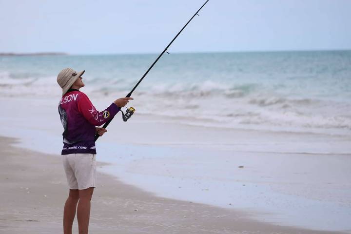 Angler wearing a wide-brim hat casts a fishing rod from a quiet sandy beach toward turquoise sea.