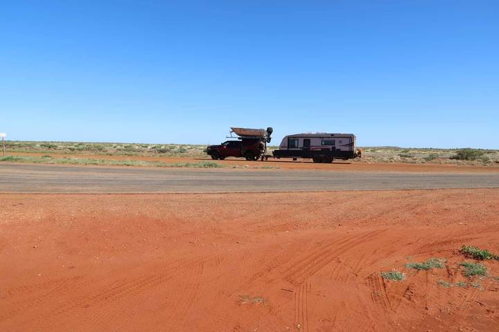 Pickup truck towing a caravan parked on a red-dust roadside in the Australian outback under a clear blue sky.