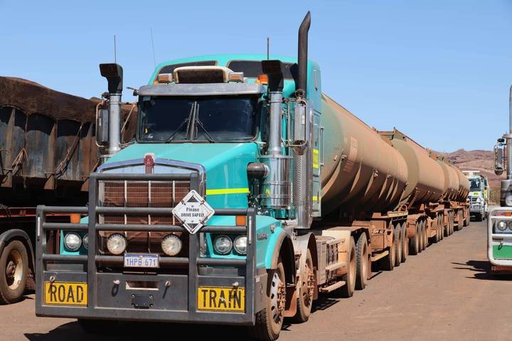 Long turquoise road-train truck with tanker trailers lined up at an outback depot under bright sun.