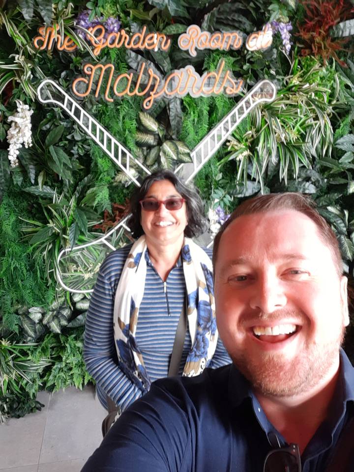 Close-up selfie of two smiling travellers against a lush indoor wall of green plants.