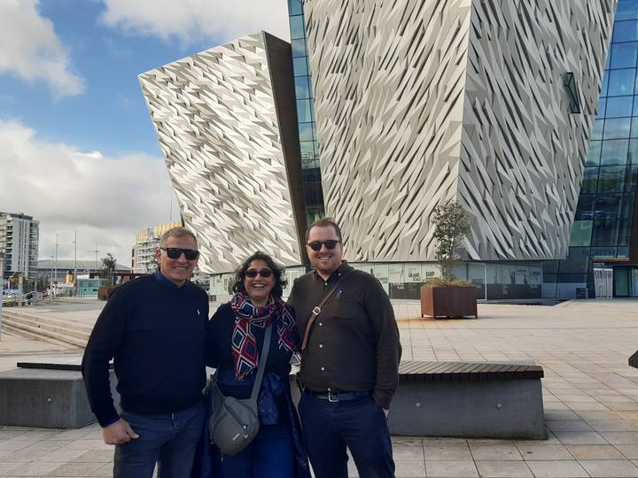 Three travellers pose in front of the angular façade of Titanic Belfast on a bright day.