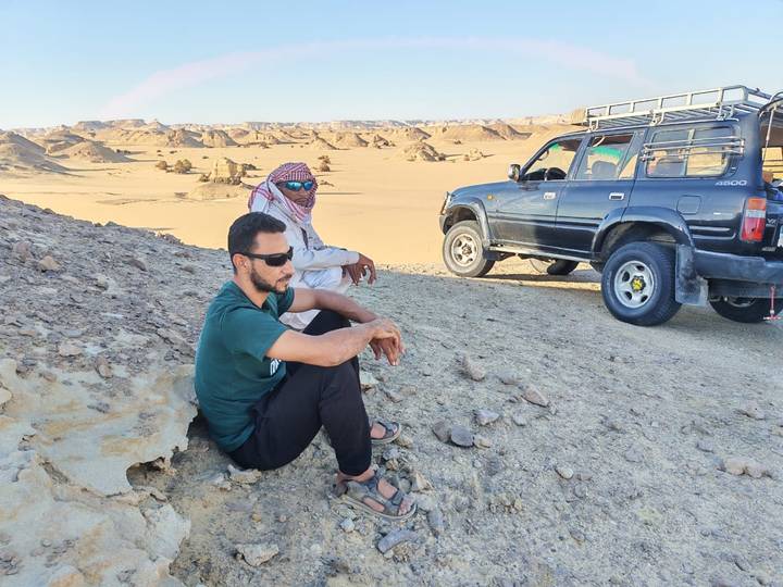 Two men rest beside an off-road vehicle amid Egypt’s sandy desert hills under bright sunlight.