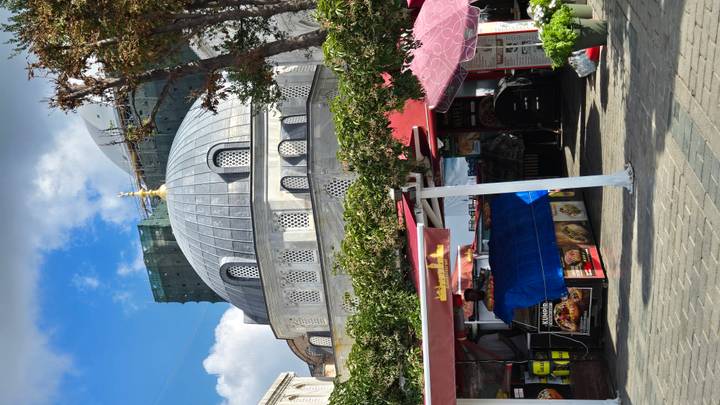 A grey–domed Ottoman-style mosque framed by small street food stalls and bright blue sky with scattered clouds.