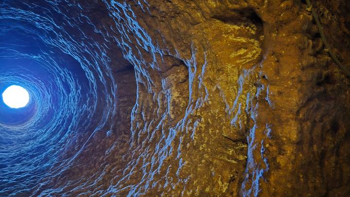 View looking up a rugged cylindrical shaft of an underground cave illuminated by daylight from a small opening above.