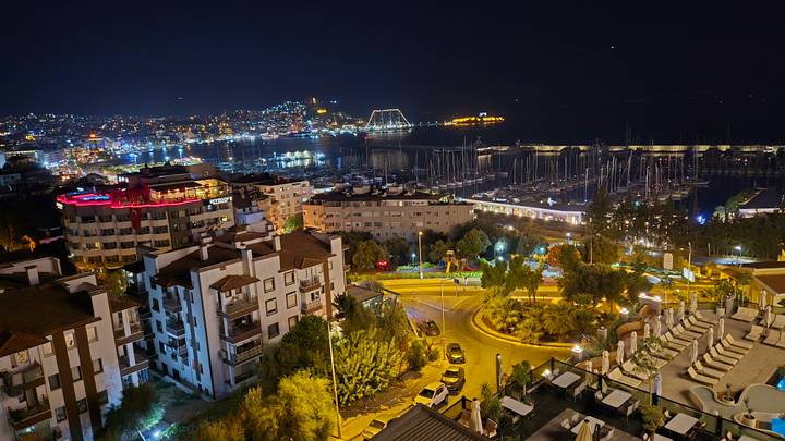 Nighttime panorama of a coastal town with illuminated marina, yacht masts and hillside lights.