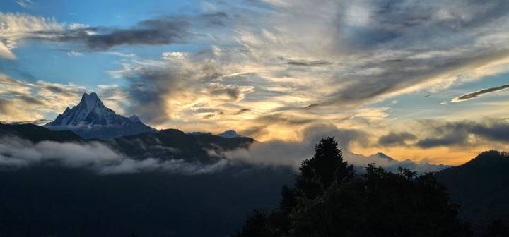 Dramatic sunrise clouds swirl above snow-capped Himalayan peaks as mist drifts through a deep valley.