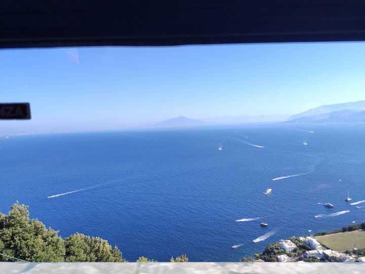 Expansive view over the deep-blue Bay of Naples with distant Mount Vesuvius, seen through a vehicle window.