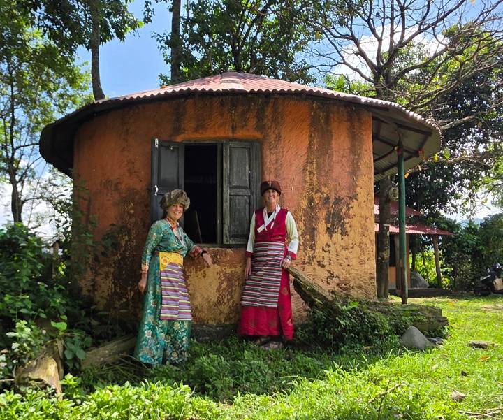 Two women in colorful traditional dress stand outside a rustic circular mud house surrounded by lush greenery.