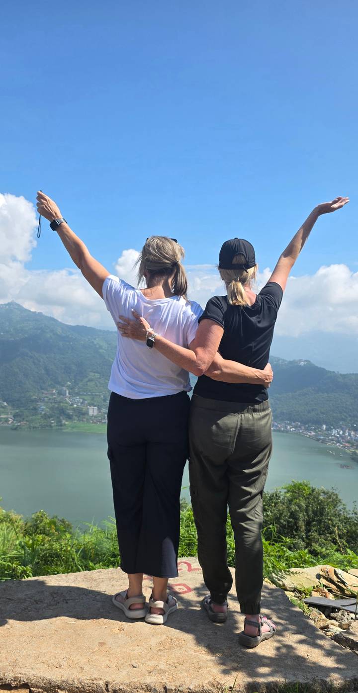 Two women embrace with arms raised atop a viewpoint overlooking a wide lake and green hills.
