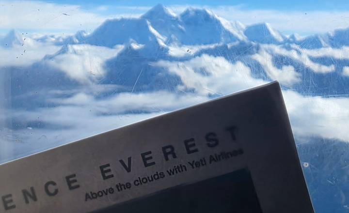View of snowy Himalayan peaks through a scratched plane window with an Everest flight brochure in the foreground.
