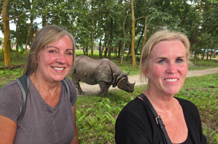 Two smiling women stand in front of a grazing one-horned rhinoceros in a lush forest reserve.