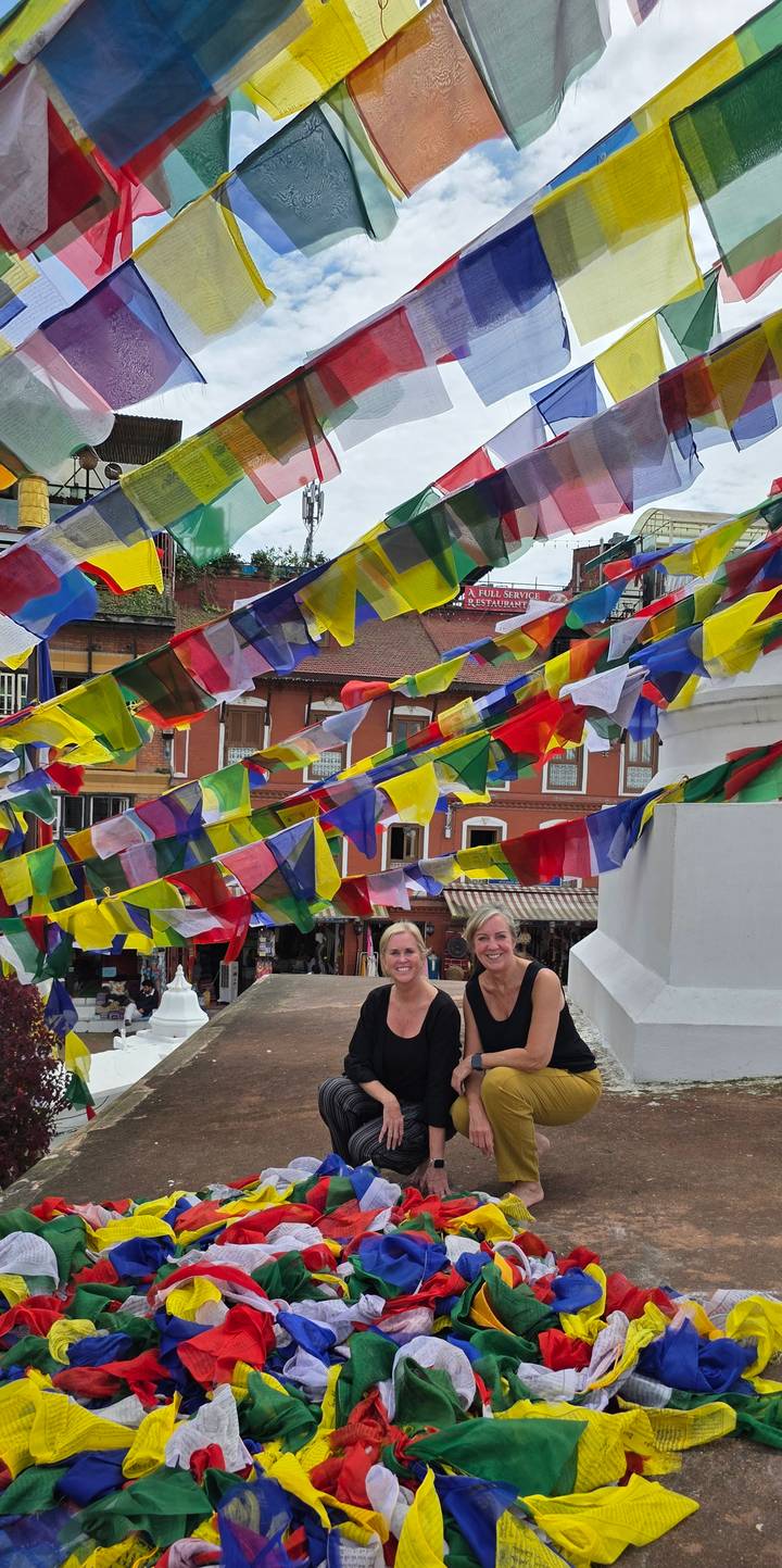 Colorful prayer flags flutter as two women smile beneath them in a vibrant Kathmandu street scene.