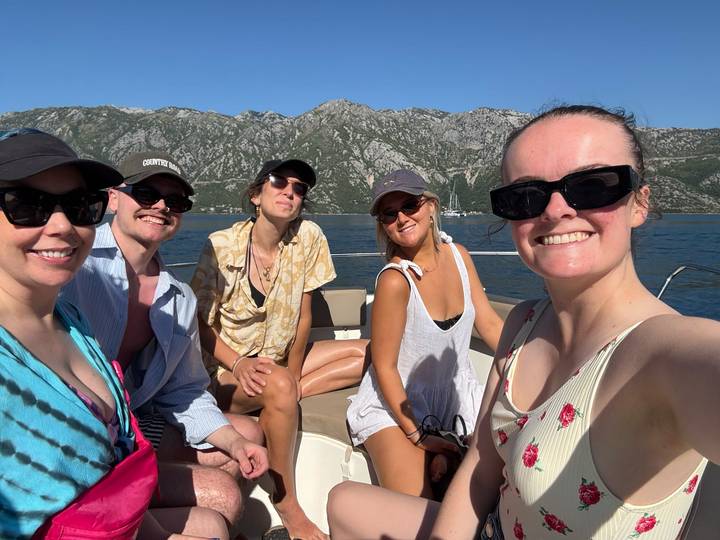 Five friends smile on a small boat with rocky mountains rising behind a clear blue bay.