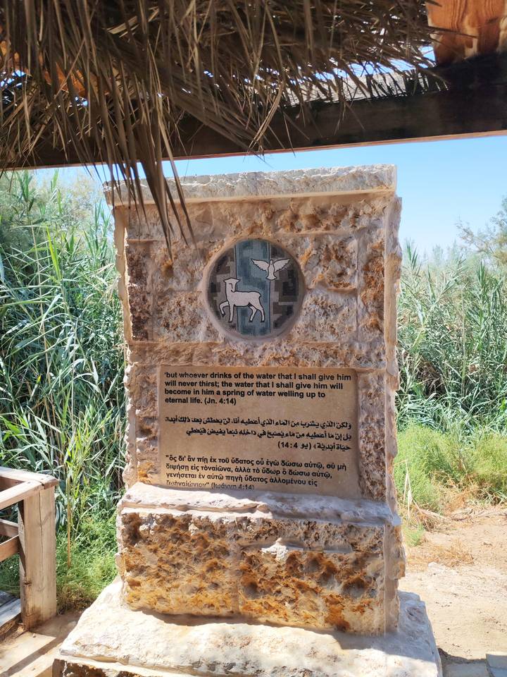 Stone plaque featuring a mosaic of a lamb and dove above a multilingual biblical verse, set outdoors among greenery.