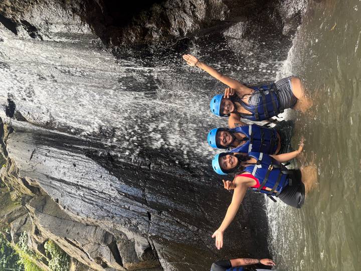 Three adventurers in helmets and life jackets celebrating beneath a cascading jungle waterfall.