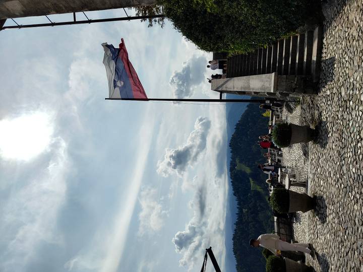 Stone terrace with tables and visitors beneath a flying Slovenian flag, overlooking forested hills and a distant lake.