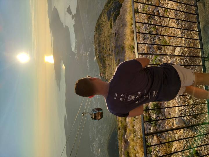Traveler looking toward a golden sunset over a bay and islands, with cable-car gondolas gliding below the viewpoint.