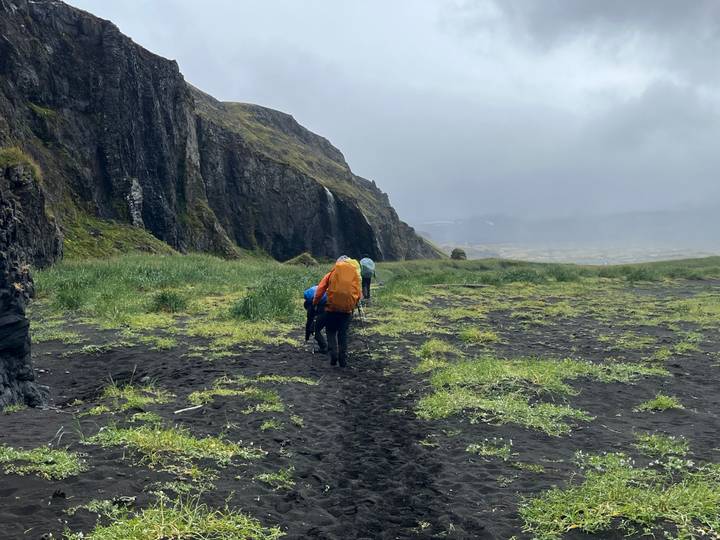 Backpackers trekking across a black-sand valley beneath towering volcanic cliffs on a misty day.