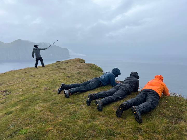 Guide pointing out to sea while companions lie prone at the edge of a grassy cliff in drizzly weather.