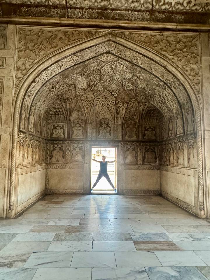 Person poses star-shaped in ornate Mughal interior chamber with intricate stucco ceiling patterns.