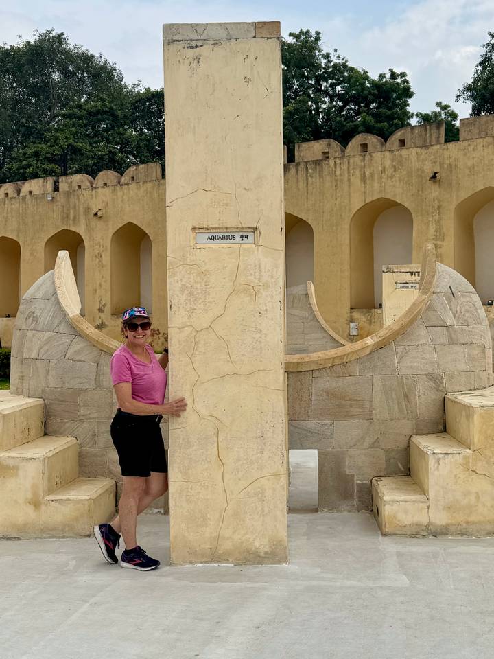 Smiling woman stands beside an engraved zodiac pillar marked 'Aquarius' at Jantar Mantar observatory.