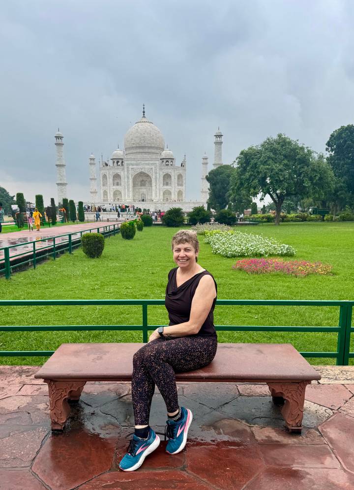 Woman smiles on a bench in lush Mughal gardens with the Taj Mahal rising majestically behind.