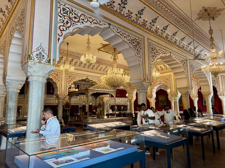 Opulent interior of Jaipur City Palace museum with chandeliers, ornate arches and visitors examining exhibits.