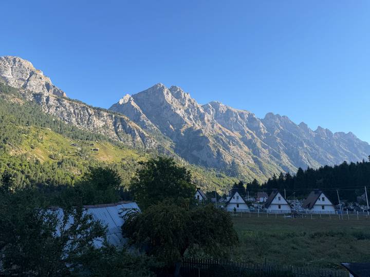 Sharp alpine peaks tower over a small village of A-frame houses in a lush valley under a clear blue sky.