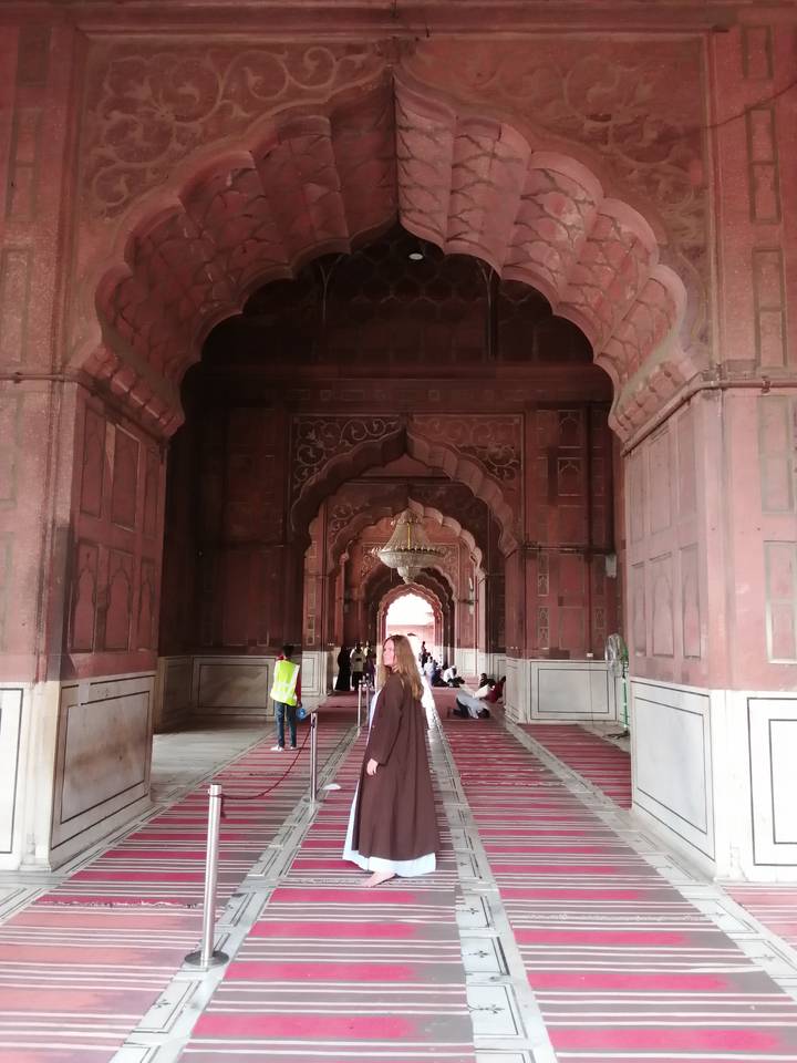 Interior arcade of red sandstone Mughal architecture with hanging chandelier and visitors seated on carpets.