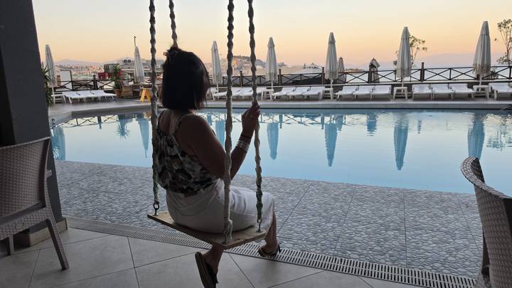 Woman on swing overlooking hotel pool at tranquil sunset with umbrellas and water reflections.