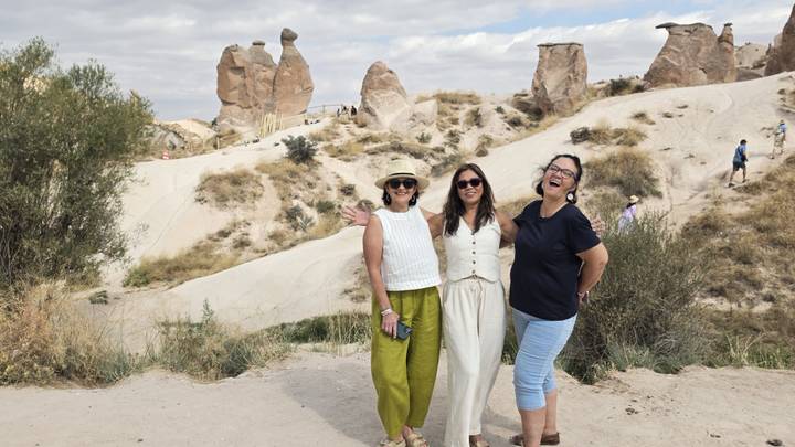 Three women pose smiling in front of Cappadocia’s distinctive fairy-chimney rock formations under a partly cloudy sky.