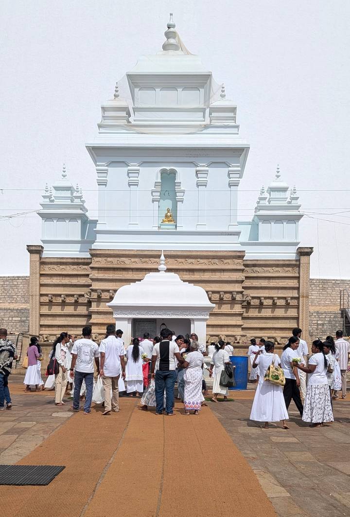 Crowds gather in white clothing at a bright Buddhist shrine with layered stupa-like towers.