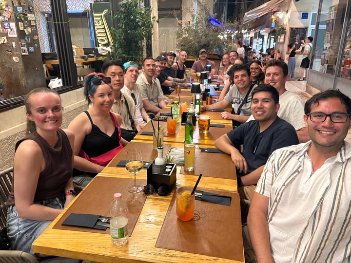 Long table of travellers sharing food and drinks inside a cosy restaurant setting.