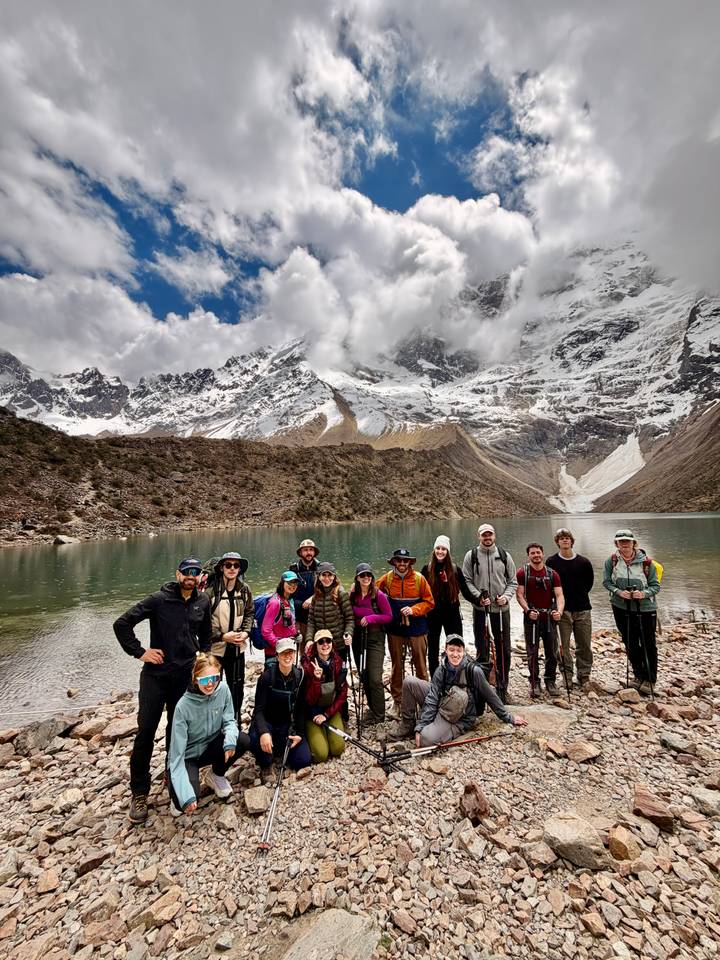 Hiking group posing beside a turquoise alpine lake backed by snowy Andean peaks and dramatic clouds.