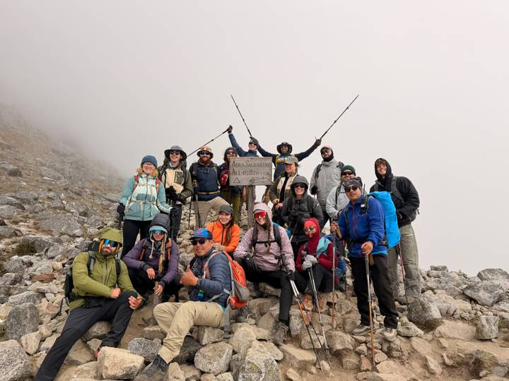 Trekking team celebrating at misty mountain pass, holding a wooden sign noting altitude.