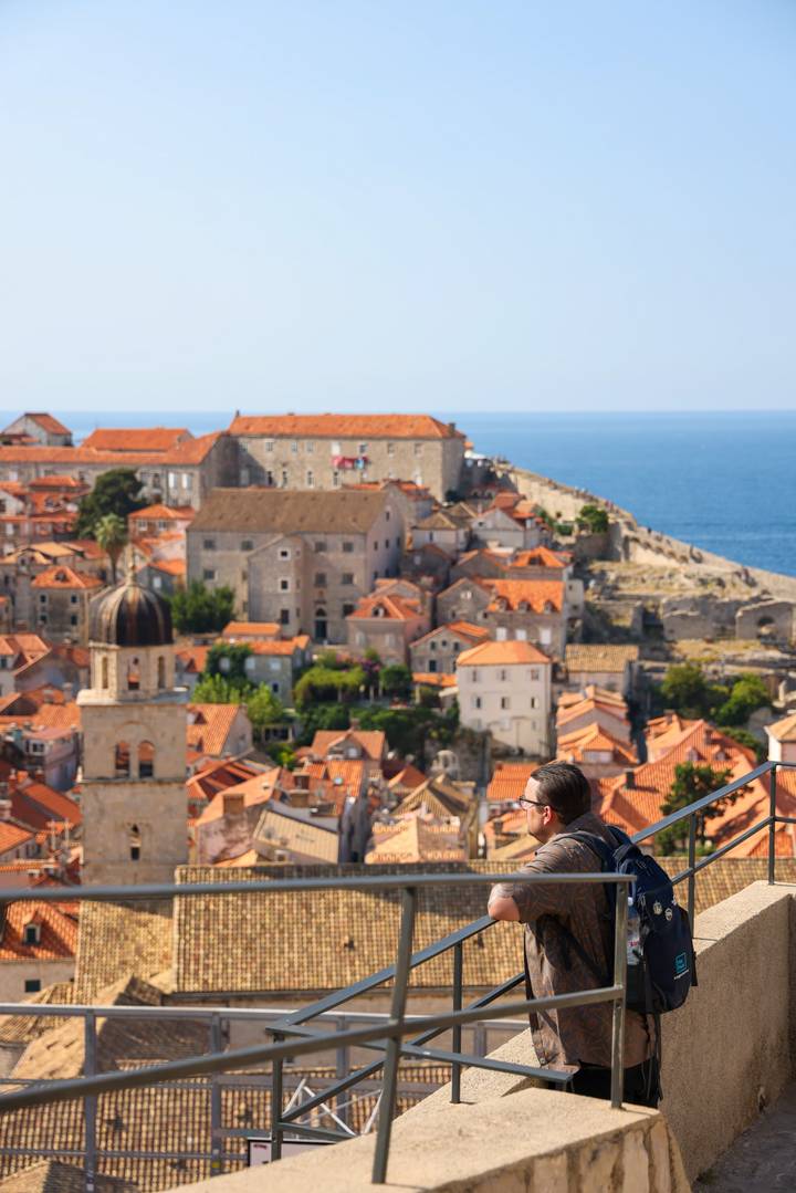 Blurred foreground figure with panoramic view over Dubrovnik’s terracotta rooftops and Adriatic Sea.