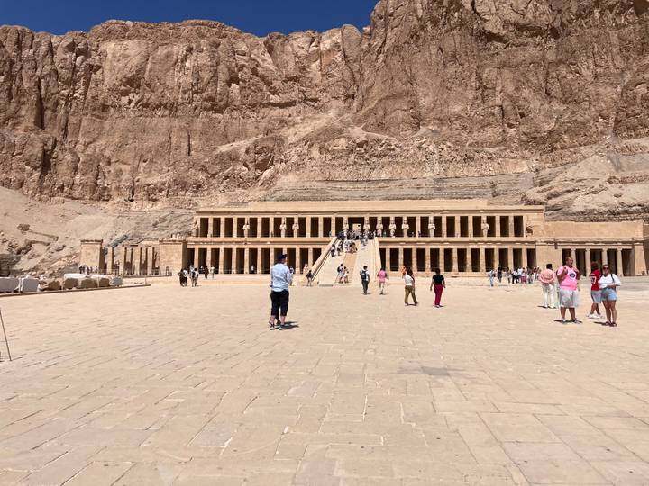 Terraced façade of Hatshepsut Temple against towering desert cliffs with tourists exploring the courtyard.